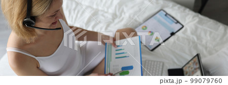 Woman in headphones holds charts sits on bed and holds online conference 99079760
