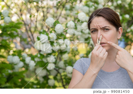 A woman suffers from a clothespin on her nose while walking in a flowery park. Allergy. A woman suffers from a clothespin on her nose while walking in a flowery park. Allergy. 99081450