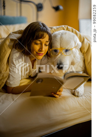 Woman with her cute dog reading book in bed Woman with her cute dog reading book in bed 99082259