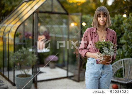 Woman with flowerpot in garden with greenhouse behind 99083526