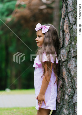 A beautiful girl in a pink dress stands near a tree in the forest and dreams 99084484
