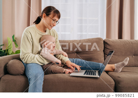 Young mom in glasses using laptop while sitting on couch with her baby at home Young mom in glasses using laptop while sitting on couch with her baby at home 99085821
