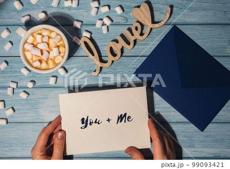 YOU plus ME text on valentine card inscription positive quote phrase Female hands holding greeting card blue envelope with white cup of coffee and marshmallows on wooden blue background. YOU plus ME text on valentine card inscription positive quote phrase Female hands holding greeting card blue envelope with white cup of coffee and marshmallows on wooden blue background. 99093421