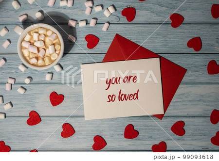 YOU ARE SO LOVED text on valentine postcard inscription positive quote phrase Greeting card with red envelope with white cup of coffee and marshmallows on wooden background. Romantic Small hearts YOU ARE SO LOVED text on valentine postcard inscription positive quote phrase Greeting card with red envelope with white cup of coffee and marshmallows on wooden background. Romantic Small hearts 99093618