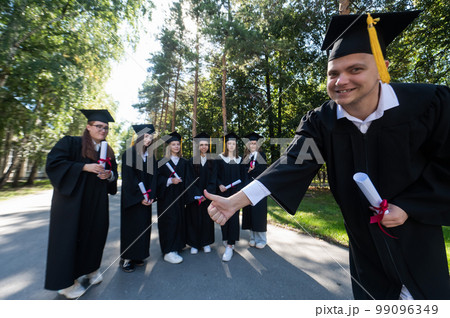 Happy young caucasian male graduate showing thumbs up. A group of graduate students outdoors. Happy young caucasian male graduate showing thumbs up. A group of graduate students outdoors. 99096349