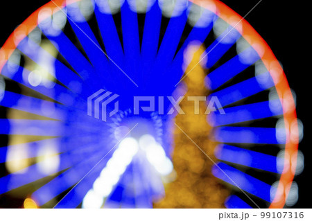 Ferris wheel decorated with blue illumination and a large Christmas tree decorated with yellow illumination on a black background at night. Beautiful New Year and Christmas holiday blurred background 99107316