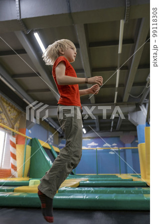 Blonde teenage boy jumps on trampoline in an entertainment center for children. Vertical frame. Blonde teenage boy jumps on trampoline in an entertainment center for children. Vertical frame. 99118308