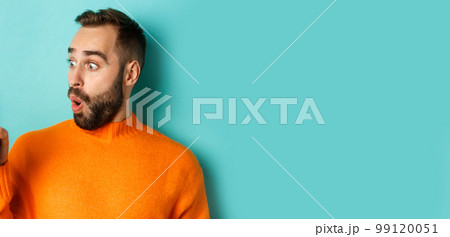 Close-up of young bearded man showing phone screen and looking amazed, wearing orange sweater, standing against studio background Close-up of young bearded man showing phone screen and looking amazed, wearing orange sweater, standing against studio background 99120051