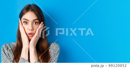 Close-up portrait of surprised cute woman holding hands on cheeks and gasping, stare at camera shocked, standing against blue background Close-up portrait of surprised cute woman holding hands on cheeks and gasping, stare at camera shocked, standing against blue background 99120052