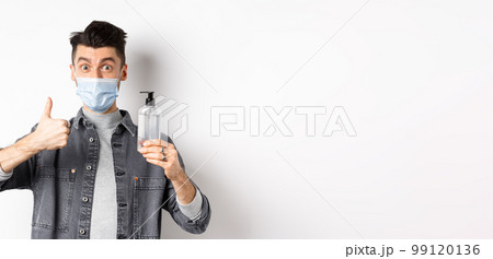 Healthy people and covid-19 concept. Excited man in sterile medical mask holding bottle of good hand sanitizer, show thumb up, recommend antiseptic, standing against white background 99120136