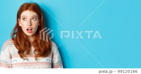 Close-up of impressed redhead girl staring left amazed, open mouth in awe and glancing at promo, standing in winter sweater against blue background 99120166