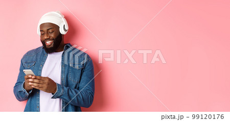 Handsome stylish Black man listening music in headphones and writing message on smartphone, smiling happy, standing over pink background Handsome stylish Black man listening music in headphones and writing message on smartphone, smiling happy, standing over pink background 99120176