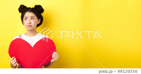 Sad and lonely asian girl standing in make-up with big red heart cutout, left alone on Valentines day, looking left disappointed or upset, standing on yellow background Sad and lonely asian girl standing in make-up with big red heart cutout, left alone on Valentines day, looking left disappointed or upset, standing on yellow background 99120550