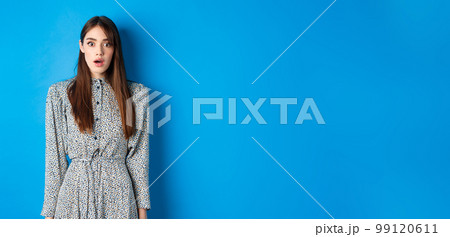 Shocked cute girl in dress looking at camera with opened mouth, gasping startled, see something shocking, standing on blue background 99120611
