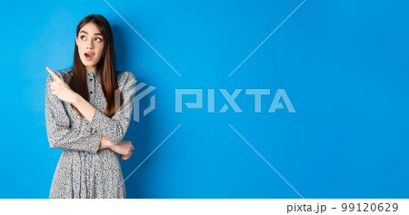 Wow look there. Excited pretty lady in dress looking and pointing left wondered, checking out advertisement, standing on blue background 99120629