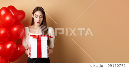 Surprised beautiful woman in romantic outfit, standing near heart balloons and looking at her gift on Valentines day, standing on beige background 99120772