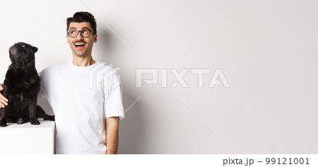 Amazed young man in glasses hugging his dog, pet owner and pug staring at upper left corner promo offer, standing over white background Amazed young man in glasses hugging his dog, pet owner and pug staring at upper left corner promo offer, standing over white background 99121001