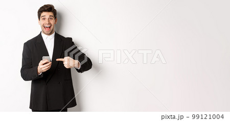 Image of cheerful businessman looking amazed, pointing at mobile phone, standing in suit over white background 99121004