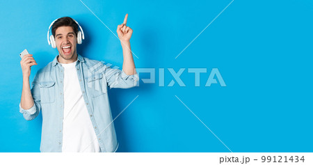 Joyful handsome man dancing with smartphone, listening music in headphones and pointing finger up, standing over blue background Joyful handsome man dancing with smartphone, listening music in headphones and pointing finger up, standing over blue background 99121434