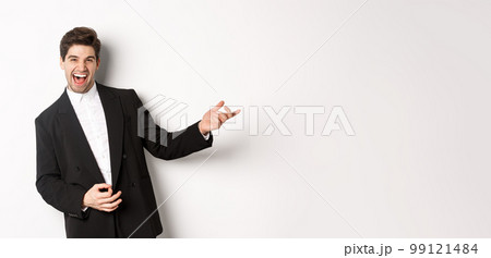 Portrait of happy man dancing at party, playing on invisible guitar and laughing, standing in black suit against white background 99121484