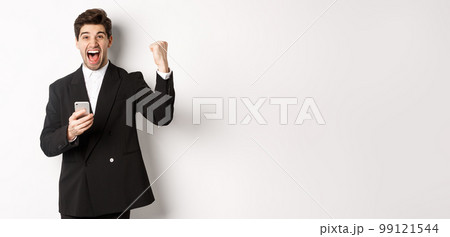 Portrait of happy handsome man in suit, rejoicing, achieve goal on mobile app, raising fist up and shouting yes, holding smartphone, standing against white background 99121544