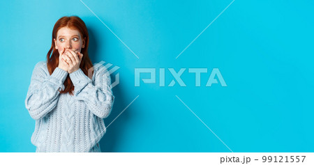 Shocked and anxious redhead girl staring left scared, covering mouth and gasping, standing over blue background in sweater Shocked and anxious redhead girl staring left scared, covering mouth and gasping, standing over blue background in sweater 99121557