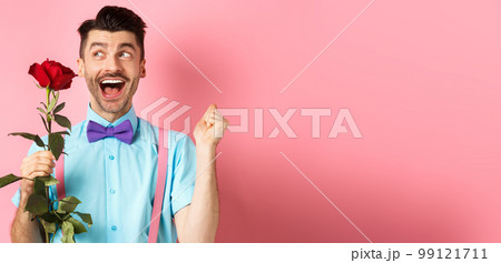 Romance and Valentines day concept. Cheerful man in bow-tie screaming from happiness, holding red rose and jumping on date, standing over pink background 99121711