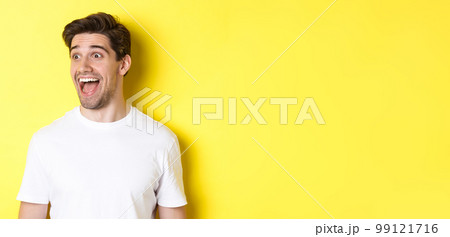 Image of happy man checking out promo, looking left with amazement, standing in white t-shirt against yellow background Image of happy man checking out promo, looking left with amazement, standing in white t-shirt against yellow background 99121716