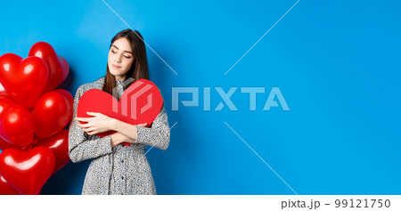 Valentines day. Romantic pretty woman in dress hugging big red heart cutout and looking dreamy, thinking of love, standing on blue background Valentines day. Romantic pretty woman in dress hugging big red heart cutout and looking dreamy, thinking of love, standing on blue background 99121750