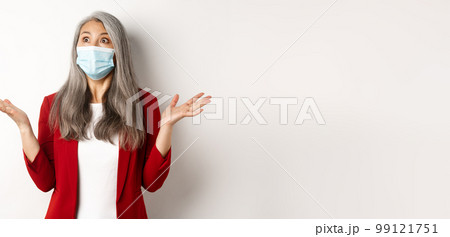 Coronavirus and business concept. Surprised asian businesswoman in face mask, spread hands sideways and staring shocked at upper left corner, white background 99121751