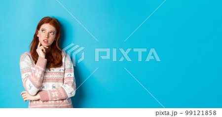 Image of thoughtful redhead girl looking at upper left corner, thinking or making choice, standing in winter sweater over blue background 99121858