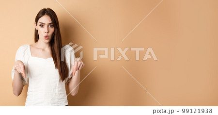 Wow amazing. Impressed young woman look astonished by product, pointing fingers down at logo, standing on beige background 99121938