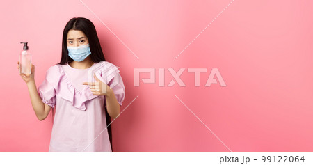 Healthy people and covid-19 pandemic concept. Skeptical asian woman frowning, wearing face mask, pointing at bottle of hand sanitizer, standing against pink background 99122064