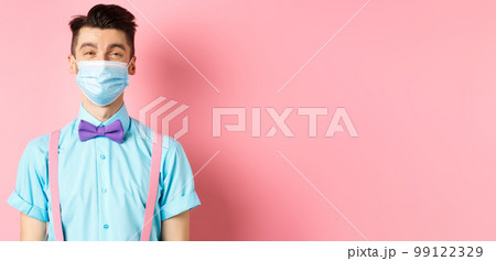Covid-19, pandemic and health concept. Portrait of smiling man in medical mask feeling happy, standing in festive bow-tie and suspenders, pink background Covid-19, pandemic and health concept. Portrait of smiling man in medical mask feeling happy, standing in festive bow-tie and suspenders, pink background 99122329
