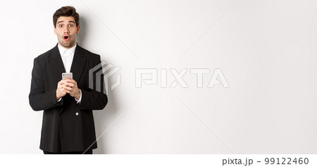 Amazed handsome guy in black suit reacting to cool promo offer, holding mobile phone, standing against white background Amazed handsome guy in black suit reacting to cool promo offer, holding mobile phone, standing against white background 99122460