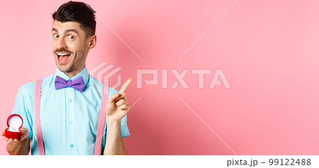 Valentines day. Funny guy with moustache and bow-tie, showing engagement ring and pointing finger at upper right corner, standing on pink background Valentines day. Funny guy with moustache and bow-tie, showing engagement ring and pointing finger at upper right corner, standing on pink background 99122488