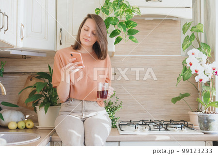 woman is sitting on table in kitchen chatting in a smartphone and drinking coffee brewing pourover 99123233
