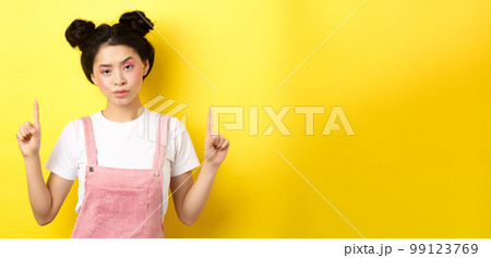 Annoyed and tired asian girl with pink glamour makeup, pointing fingers up and look bothered, standing on yellow background Annoyed and tired asian girl with pink glamour makeup, pointing fingers up and look bothered, standing on yellow background 99123769