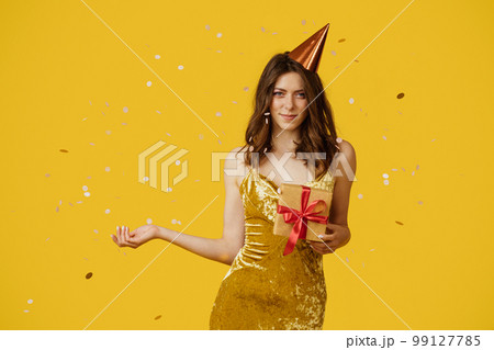Happy lady holding wrapped gift boxes, wearing festive dress and birthday hat, yellow background with falling confetti 99127785