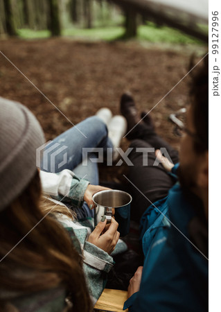 Young european guy and lady tourists in jackets rest in autumn forest, enjoy cup of hot drink, free time in camping Young european guy and lady tourists in jackets rest in autumn forest, enjoy cup of hot drink, free time in camping 99127996