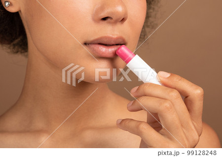 Serious young black curly lady with perfect skin applies lipstick on lips for moisturizing 99128248