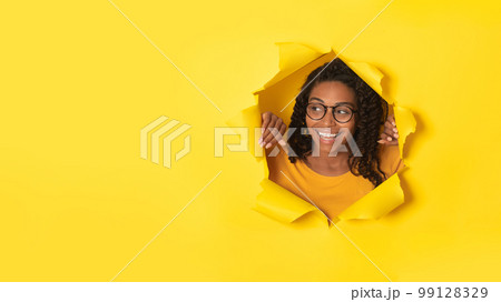 Excited african american lady in glasses posing in torn paper, looking at free space through hole on yellow background Excited african american lady in glasses posing in torn paper, looking at free space through hole on yellow background 99128329