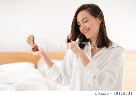 Cheerful pretty caucasian millennial lady sits on bed, combs her hair and looks at tips, enjoy good morning 99128331