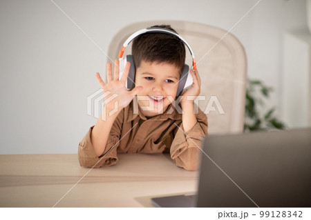 Glad european small kid in headphones waving hand, has video call with teacher, tutor at table 99128342