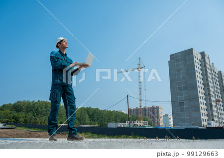 A male builder in a helmet stands against the background of a construction site with a laptop in his hands.  99129663