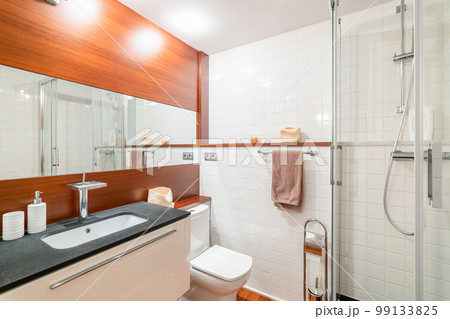 Bathroom with one wall of deep caramel wood paneling and large mirror between them. In contrast, white sink is built into black marble countertop. Mirror reflects shower cabin with glass doors. Bathroom with one wall of deep caramel wood paneling and large mirror between them. In contrast, white sink is built into black marble countertop. Mirror reflects shower cabin with glass doors. 99133825