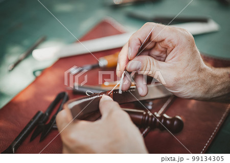 closeup hand working process leather handcraft in the leather workshop. Man holding crafting tool and working. He is sewing hammer to make a walet. Tanner in old tannery. 99134305