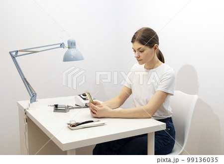 Nail beautician hold gadget, looking at smartphone, sitting at workstation in nail beauty treatment salon. Professional tools are on white table. Studying manicurist. Closeup, horizontal 99136035