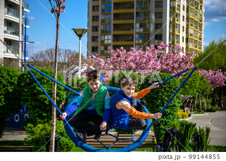 Cheerful boy with his toddler brother swinging in the swing-nest. Entertainment for preschoolers outdoors. Relaxation friendship concept Cheerful boy with his toddler brother swinging in the swing-nest. Entertainment for preschoolers outdoors. Relaxation friendship concept 99144855