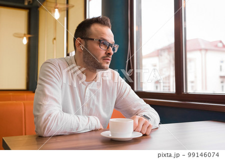 Young attractive businessman in a white shirt, glasses and headphones drinks coffee and looks thoughtfully out the window 99146074